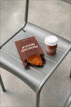 Coffee cup mockup and paper bag mockup styled on a café chair with a croissant. Paper bag mockup and coffee cup mockup together for a coffee shop branding scene. Coffee cup mockup, paper bag mockup, packaging mockup, coffee shop mockup, bakery mockup, café mockup, high quality mockup, PSD file.
