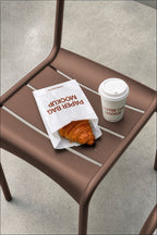 Coffee cup mockup and paper bag mockup styled on a café chair with a croissant. Paper bag mockup and coffee cup mockup together for a coffee shop branding scene. Coffee cup mockup, paper bag mockup, packaging mockup, coffee shop mockup, bakery mockup, café mockup, high quality mockup, PSD file.