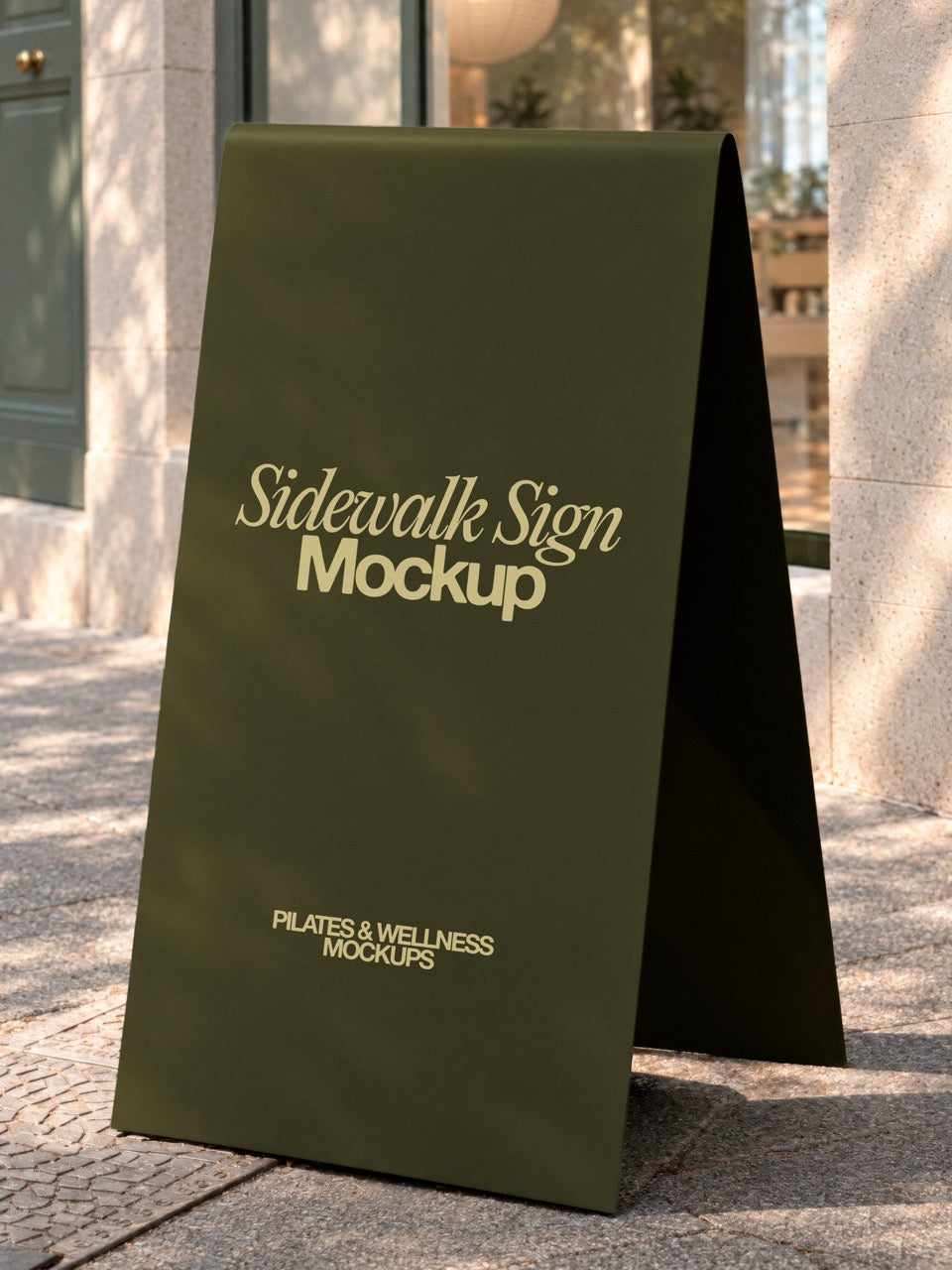 Sidewalk sign mockup with an olive A-frame board outside a stone studio entrance in dappled shade. A-frame sign mockup for pilates and wellness studios. Sidewalk sign mockup, A-frame mockup, signage mockup, studio sign mockup, pilates mockup, wellness mockup, high quality mockup, PSD file.