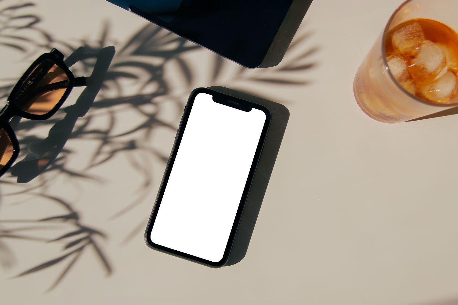 Real photo iPhone mockup PSD on sunlit desk with plant shadows, sunglasses, and iced drink