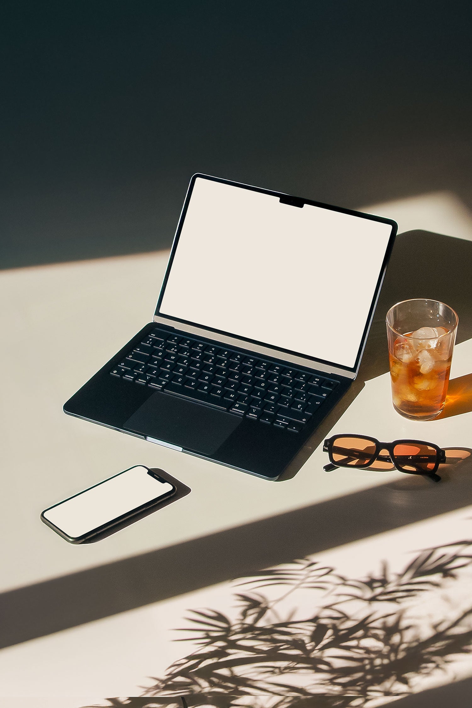 MacBook and iPhone mockup PSD on sunlit desk with plant shadows, sunglasses and iced drink
